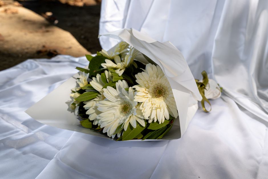 A bouquet of fresh white gerbera daisies with green foliage, wrapped in white paper and tied with a gold ribbon, is resting on a white satin cloth. The flowers are arranged in a compact, rounded style