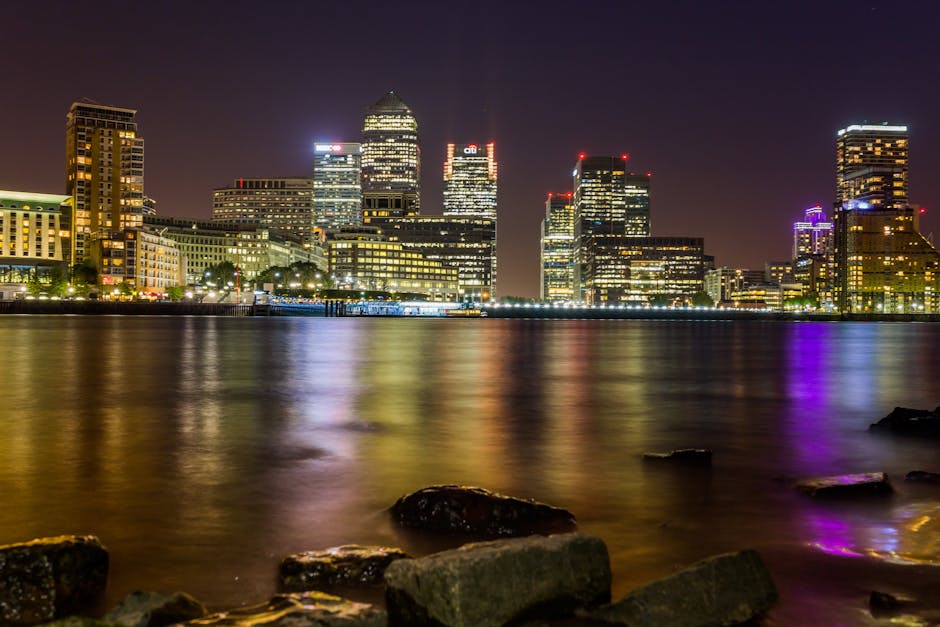 A nighttime cityscape of London's Canary Wharf area viewed from across a calm river, featuring illuminated high-rise office buildings with glass facades reflecting colorful lights onto the water. The 