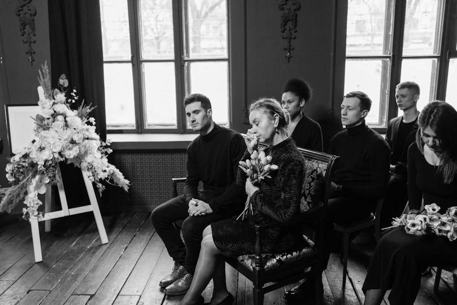 A black-and-white photograph depicting a solemn indoor scene where five individuals are seated in a row, attending a memorial or funeral service. The woman in the center, holding a bouquet of flowers,